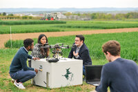Foto: Vier Personen sind auf einem Feld zu sehen, die um eine große Drohne herum stehen. Eine Person hält einen Laptop, während die anderen die Drohne betrachten und diskutieren. Im Hintergrund sind Felder und ein Traktor sichtbar.