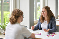 Foto: Zwei Personen sitzen an einem Tisch in der Nähe eines Fensters und unterhalten sich. Auf dem Tisch liegen Broschüren mit einem Ernährungskreis. Eine Pflanze steht auf der Fensterbank.
