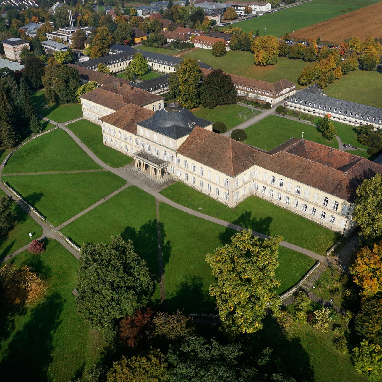 Foto: Luftaufnahme von Schloss Hohenheim, einem großen historischen Gebäude mit symmetrischem Design und Kuppeldach. Es ist von gepflegten Rasenflächen und Bäumen umgeben. Im Hintergrund sind weitere Gebäude und Felder zu sehen. Wege führen durch die Grünflächen.