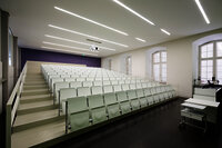 Foto: Ein moderner Hörsaal mit weißen, gestuften Sitzreihen und einem Dozentenpult. Die Wände sind hell, und die Decke hat lineare Beleuchtung. Große Fenster lassen Tageslicht herein.