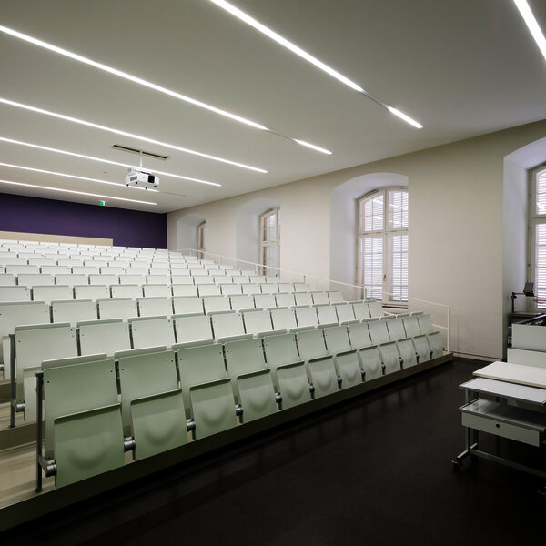 Foto: Ein moderner Hörsaal mit weißen, gestuften Sitzreihen und einem Dozentenpult. Die Wände sind hell, und die Decke hat lineare Beleuchtung. Große Fenster lassen Tageslicht herein.