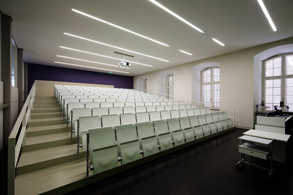 Foto: Ein moderner Hörsaal mit weißen, gestuften Sitzreihen und einem Dozentenpult. Die Wände sind hell, und die Decke hat lineare Beleuchtung. Große Fenster lassen Tageslicht herein.