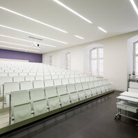 Foto: Ein moderner Hörsaal mit weißen, gestuften Sitzreihen und einem Dozentenpult. Die Wände sind hell, und die Decke hat lineare Beleuchtung. Große Fenster lassen Tageslicht herein.